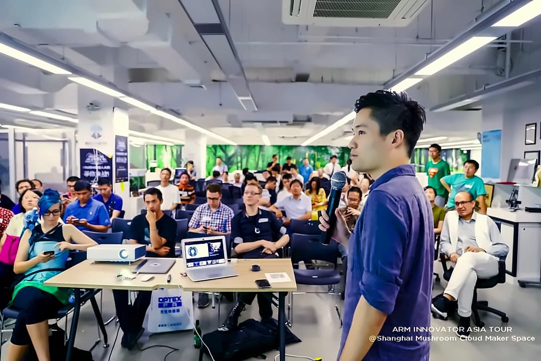 A speaker addressing a seated audience of roughly fifty developers in a bright open maker space