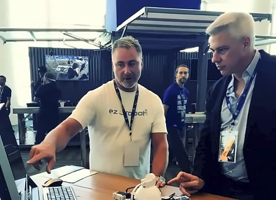 An EZ-Robot engineer in a branded shirt leaning over a desk, pointing at a screen while explaining the build to an attendee in a jacket and conference lanyard, a small white robot on the desk between them