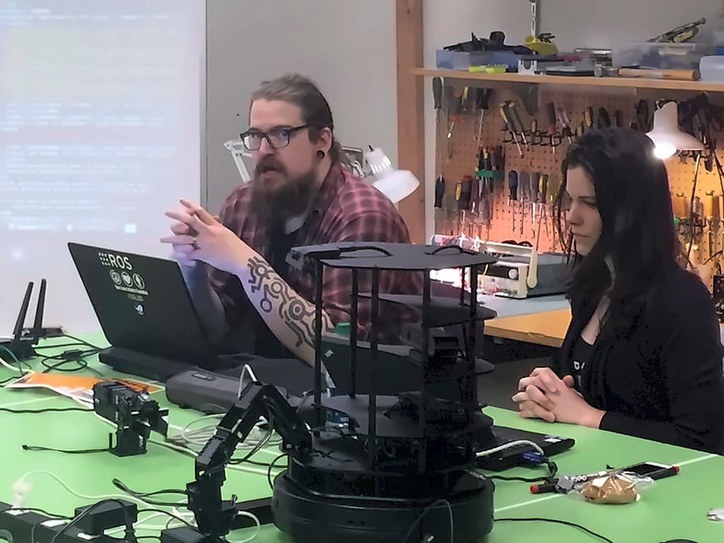 Two people at a lab bench with the robot and a laptop running ROS, a terminal of block-detection log output projected on the wall behind them