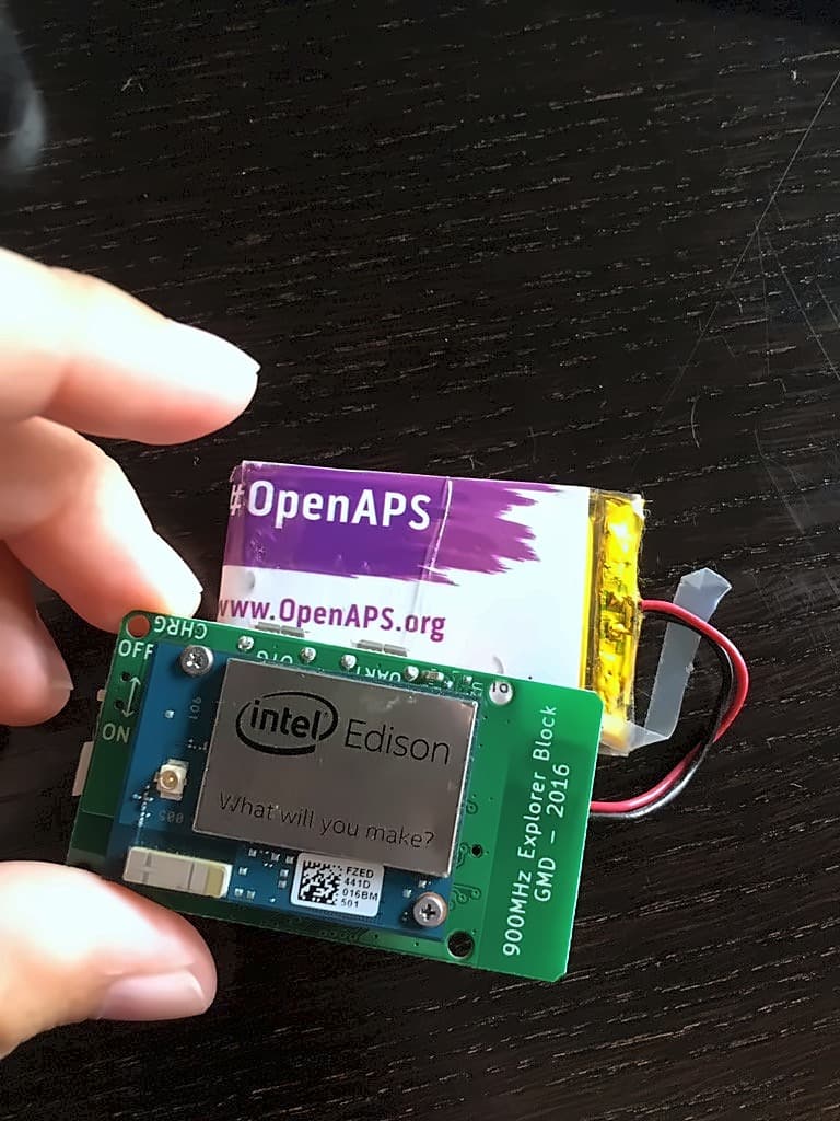 A hand holding an Intel Edison on a green 900MHz Explorer Block dated 2016, with a lithium battery taped on behind an OpenAPS sticker
