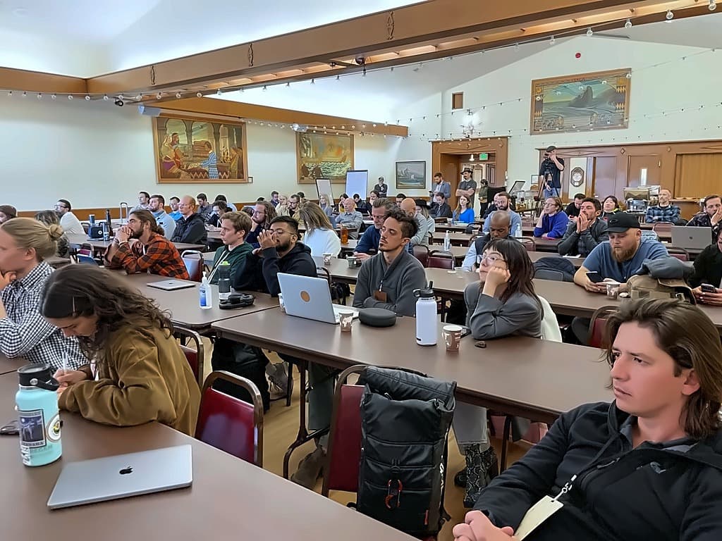 Attendees seated at long tables in a wood-beamed hall hung with murals, watching a session
