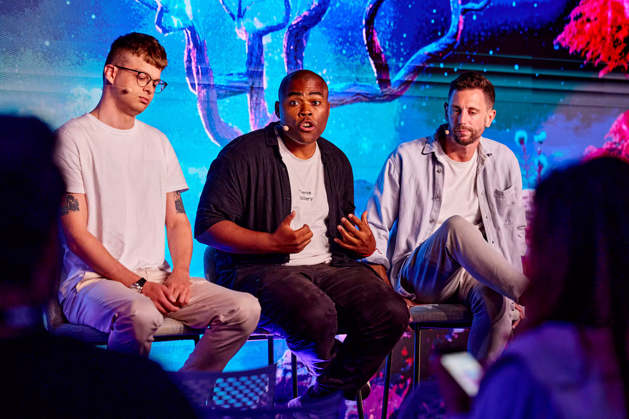 A three-person panel seated on stage, backlit by a wall-sized screen of vivid blue and magenta game art
